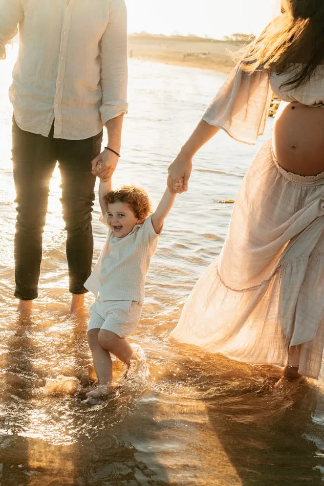 An expecting mum and dad walking their toddler through shallow water