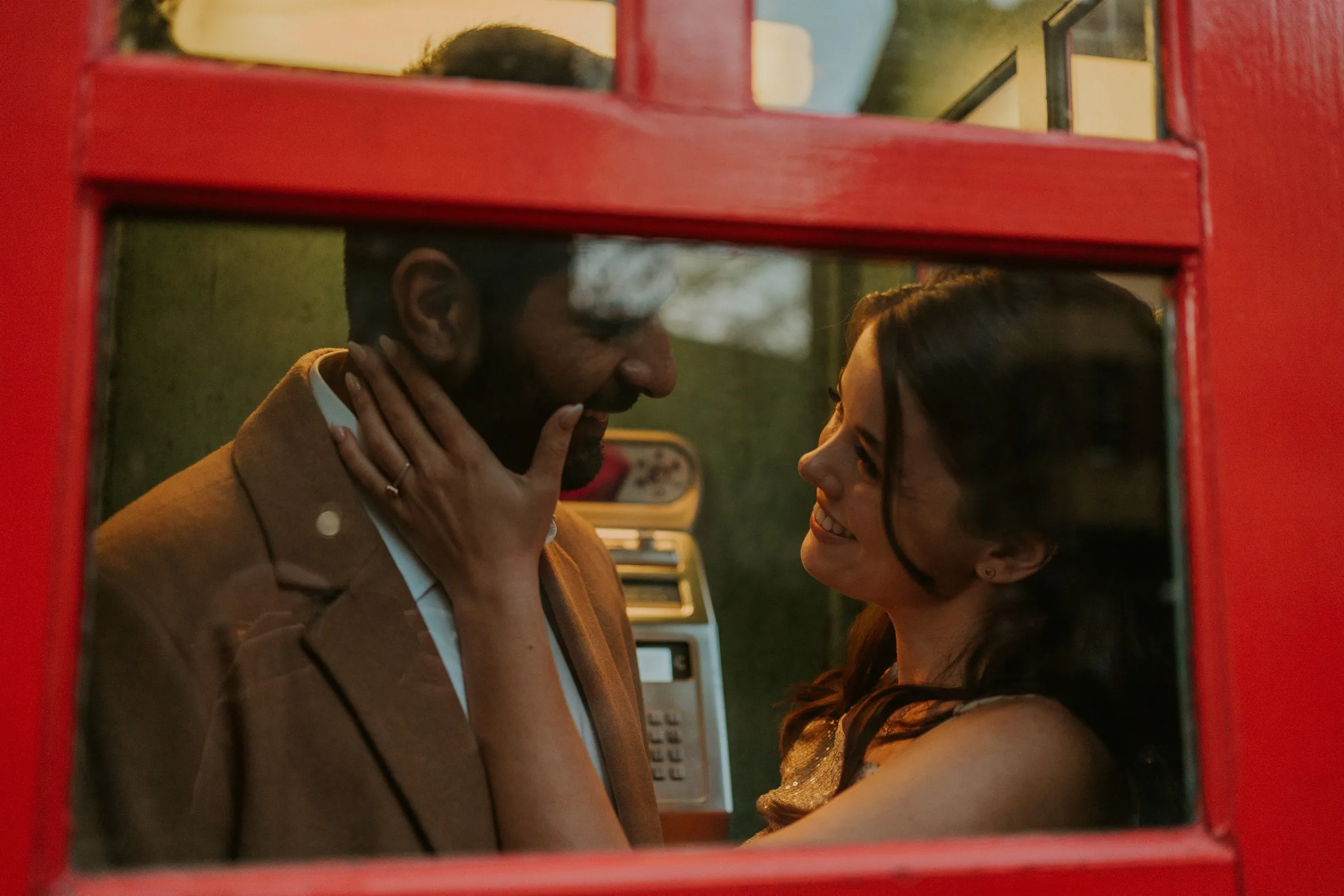 A couple smiling at each other through the glass of a red phone box