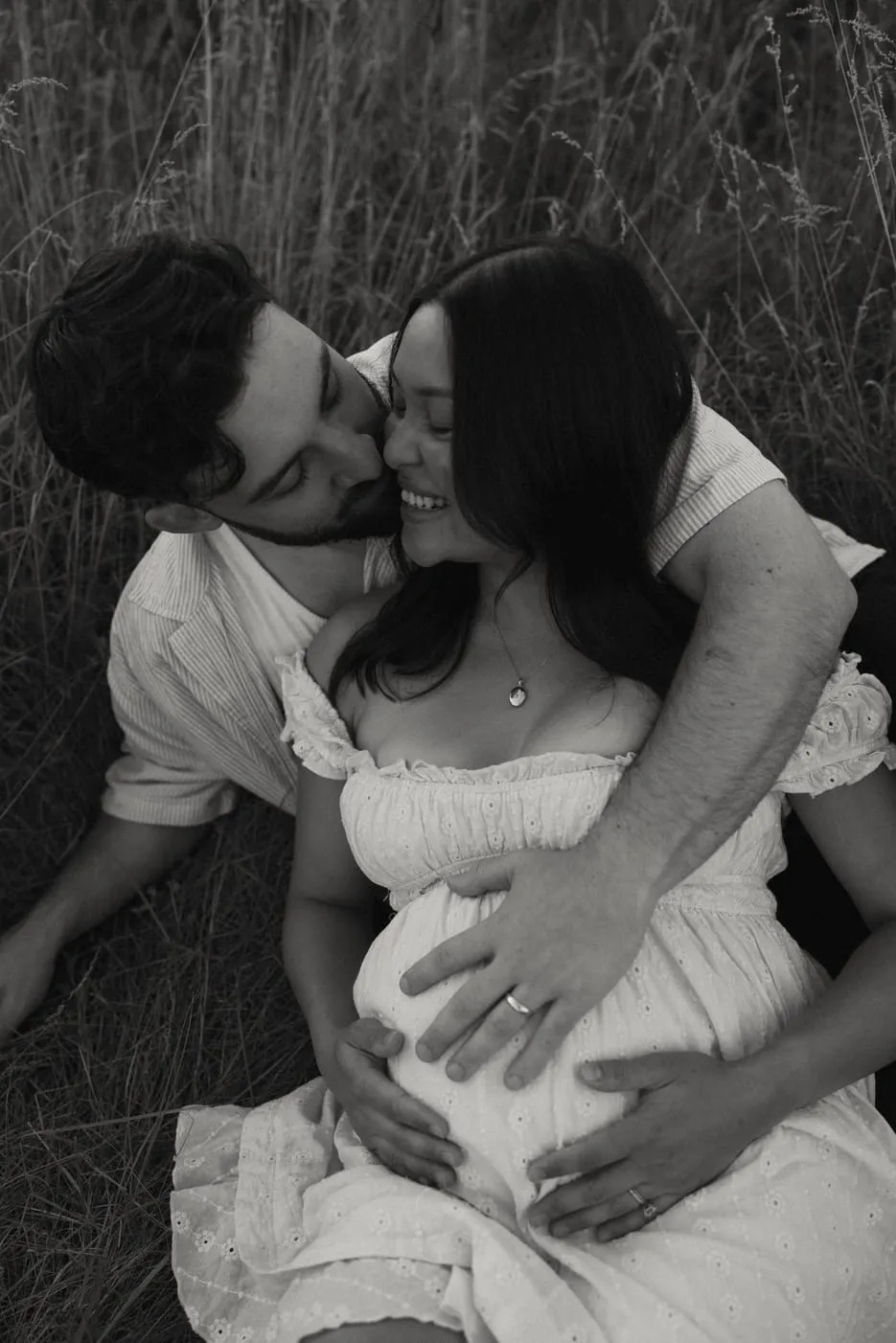 A black and white portrait of an expecting couple embracing in the grass